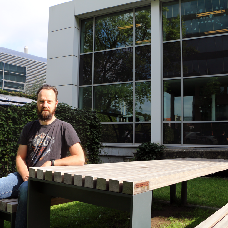 Tony zit aan een picnictafel voor het UCO-gebouw. Zijn linkerarm rust op de tafel en hij kijkt in de lens